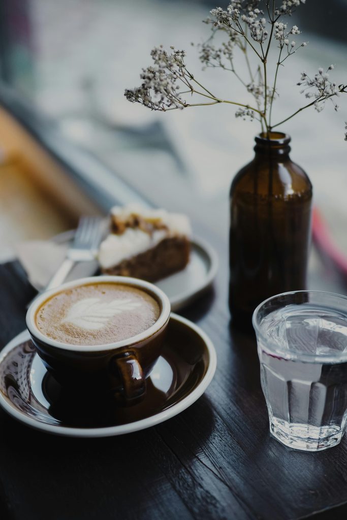 Taza de café con arte latte junto a un vaso de agua y una porción de pastel casero en una mesa de cafetería acogedora, con flores secas en un jarrón de vidrio marrón.